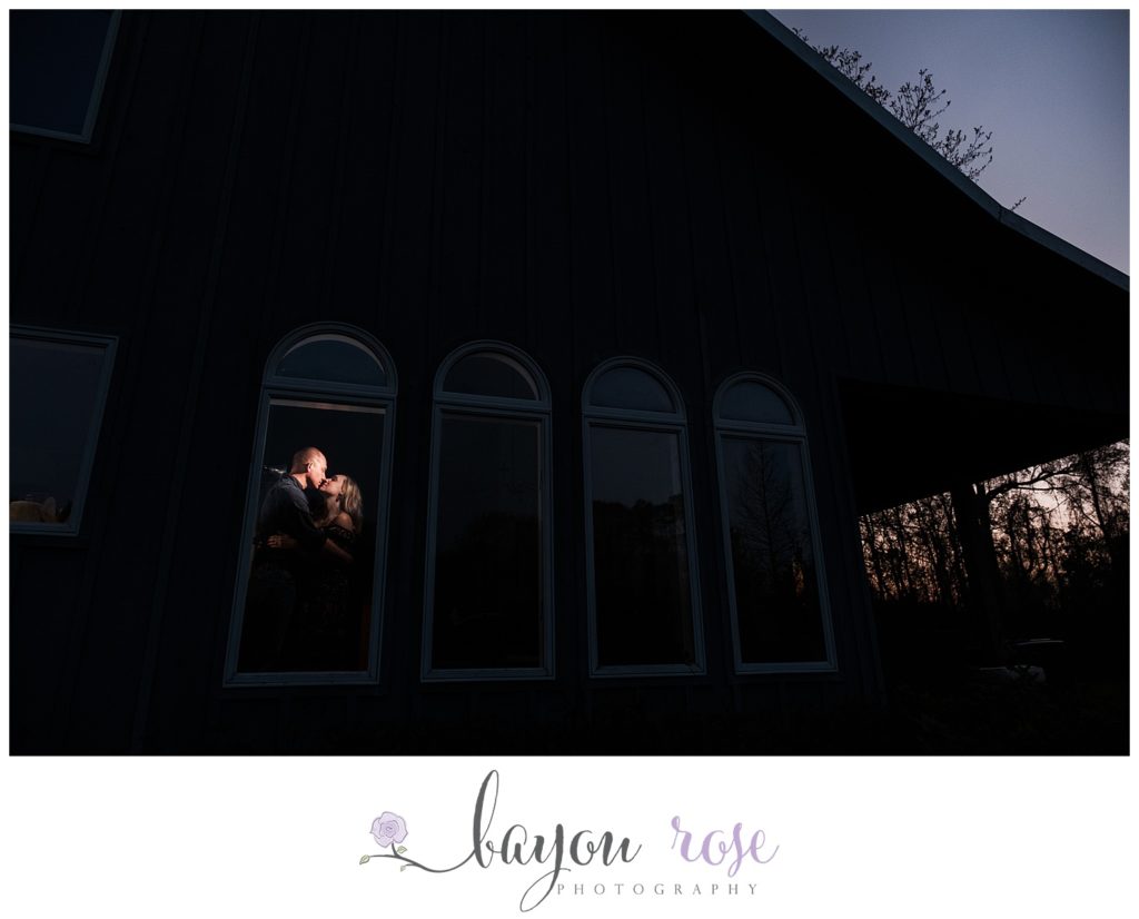 creative engagement photo of couple spotlit in window of their home as the sun sets behind them